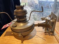 Side view of worn metal railroad caboose lamp showing base, chimney holder, metal bracket and frame on wooden table with measuring ruler next to it.