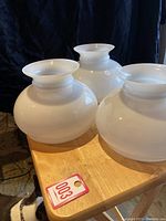Three white glass lamp globes with wide flared tops and round bulbous bases on a wooden table.