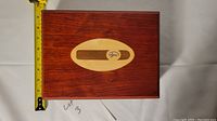 Top view of closed solid wood humidor box with oval inlay and logo on lid next to measuring tape showing width.