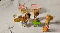 Photo showing 5 miniature metal chairs, a white and yellow kitchen cabinet, 4 miniature dolls, a green round metal table with a potted plant, and two brass-tone plastic beds with pink bedding arranged on white surface.