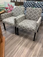 Pair of matching upholstered chairs with grey and white geometric patterned fabric and dark wood legs shown on floor.