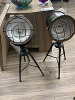 Front view showing two tripod battery operated black metal lights with protective grids on heads and extended legs.