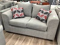 Front view of gray fabric loveseat with two red, black, gray, and white geometric pattern throw cushions on each side.