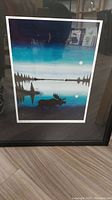 Framed art print showing a moose in silhouette by a lake with trees and moons in background under a blue sky.