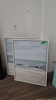 Two wood framed prints on canvas leaning against a wall, showing the beach scene with ocean and sand.