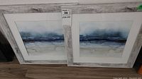 Two wood framed abstractive watercolor prints with distressed whitewashed frames, placed leaning against a wall.