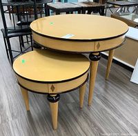 Pair of round nesting tables with light wood veneer tops, black trim edging, and tapered legs showing wood accents, placed on a laminate floor amongst other furniture in a store setting.