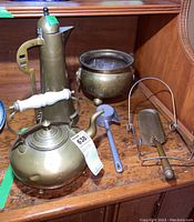 Brass coffee pot with white handle, brass kettle, footed brass bowl, and a brass plate stand with cake server including meat mallet in view on wooden shelf.