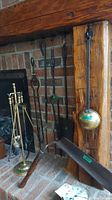 Photo showing a vintage fireplace tool set with assorted metal tools and a brass stand, leaning against a brick fireplace