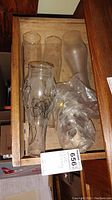 Photograph showing six glass oil lamp chimneys in a wooden drawer. Four chimneys are clear glass, two are frosted glass. One clear chimney has black floral decorations on it.
