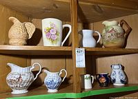 Two wooden shelves holding nine various-sized jugs and pitchers in different styles and colors, including porcelain and pottery pieces.