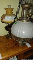 Photo showing both lamps side by side on a wooden surface. Left lamp is the amber and white floral oil lamp converted to electric. Right lamp is the white frosted globe pendant lamp with metal accents.