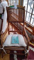 Front view of vintage wooden rocking chair with cane backrest and cushioned seat, showing attached green auction tag and book on seat.