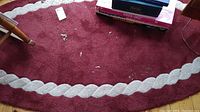Close up photo of smaller red oval rug with white scalloped border, showing some dirt and debris on surface. Includes tag marked 30.