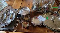 A selection of silver plated pieces including large serving trays, lidded serving dishes, small plates, and bowls arranged on a wooden table.