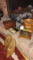 Photo shows pair of wooden shoes, vintage metal tin, wood box, ceramic bowl on wooden stand, arranged on hearth area with brick background.