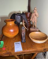 Six wooden items on a wooden shelf: elephant figurine, dog figurine, man statue, vase, bowl, candle stand.