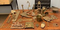 Assorted brass pieces and parts shown on a table with a black chair in the background. Includes bookends, figurines, plaques, trays, candlesticks, and decorative items. Items show tarnish, dirt, and some incomplete parts.