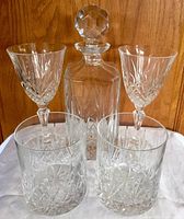 Photo showing the crystal lidded decanter with stopper and four glasses: two goblet style and two tumbler style glasses on a white cloth with wooden background.