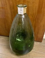 Photo showing full front view of large green glass jar with visible reflections, placed on a tiled floor against a wood panel background, tagged with seller's label.
