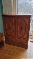 Front view of wood armoire with closed doors showing carved panel design and bronze handles.