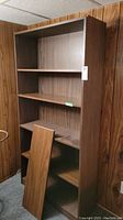 Photo front view of wooden 5-shelf bookcase with an additional detached shelf leaning inside, set against wood paneled wall.