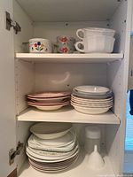 Open cabinet shelf showing various stacked ceramic plates, bowls, and cups, including plain white dishes and floral patterned pieces.
