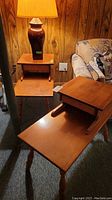 Front view of two vintage wood 2-tier end tables, one upright with a lamp on top and the other laying on its side showing the tier detail and turned legs.