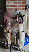 Photo showing two golf bags and several golf clubs each. One bag is white and worn with a label, the other brown and also showing wear. Golf clubs include drivers and irons.