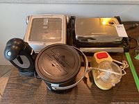 Full view of all five kitchen appliances grouped on a table: George Foreman grill, Hamilton Beach can opener, Presto deep fryer, Hamilton Beach grill, unknown black handheld appliance.