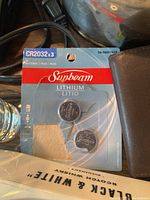 Pack of three Sunbeam CR2032 lithium batteries still in retail packaging, placed on surface with black power cord and part of brown leather-like pouch visible around it.