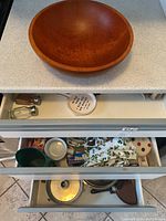 Large wooden salad bowl resting on a kitchen counter with kitchen drawers partially open showing various kitchen items including salt and pepper shakers, colander, kitchen towels, and linens.