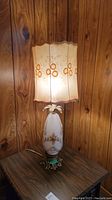 Photo of the vintage glass-based table lamp on a wood grain table against a wood paneled wall, showing the lamp lit with a floral fabric shade and detailed painted glass base with metal accents.