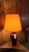 First ceramic lamp on wooden triangular side table against wood panel wall, orange pleated shade lit
