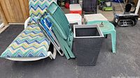 Side view of outdoor lounge chair cushion with chevron pattern along with four folding lawn chairs in green, white, and blue shades, and three small tables in green, white, and beige colors, plus black wicker-style planter.