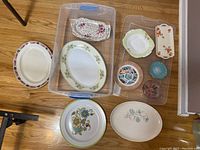 Photo showing eleven various ceramic and porcelain platters and plates in plastic bins and on floor, including oval, round, and rectangular shapes with floral patterns in multiple colors.