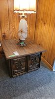 Vintage wood square end table with storage, detailed carved front panels, and textured wood grain top, shown with a lamp on top in a basement setting.