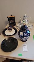 Photo of the entire set including black and gold teapot and tray, white and blue floral teapot and tray, small blue and gold teapot on plate, black swirl plate, and black and white floral ginger jar