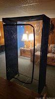 Front and side view of black portable fabric wardrobe with clear plastic front zipped open, set in a wood-paneled basement room.