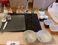 Photo showing assortment of kitchen items including heavyweight cutting boards, glassware, bowls, salt and pepper shakers, and small containers.