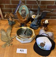 Multiple eagle figurines, decorative plate with eagle image, eagle-shaped candle, eagle-themed trucker hats displayed on a wooden surface with a brick wall background.