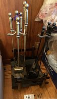 Two fireplace tool sets and a metal log holder placed next to each other on a wooden floor against a wood-paneled wall.