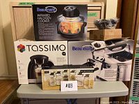 Photo of boxed Beaumark Infrared Halogen Cooker, Beaumark Stainless Steel Deep Fryer, Tassimo Coffee Machine, spice rack, and tortilla makers on a table.