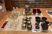 Lot showing multiple goblets of silver plated brass and brass, small clear glass vinegar bottles, retro red dessert glasses, a silver plated pitcher, and small drinking glasses laid out on table.