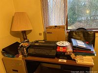 Photo showing a manual black typewriter on a desk alongside boxes of envelopes, folders, a briefcase bag, and a Dell cardboard box. The setting is next to a window with outdoor view.