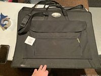 Full view of black fabric art carrying bag with shoulder strap, front zippered and flap pockets, and carrying handles.