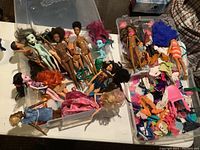 Wide view of various Barbie and fashion dolls with accessories in plastic bins on a table