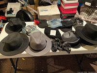 Full view of nine hats arranged on table. Various styles and materials visible including fedoras, woven hats, embellished denim, leather hat