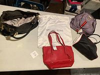 Overview of four purses on table: black MK chain bag, red MK tote with dust bag, lavender satchel, Prada black tote