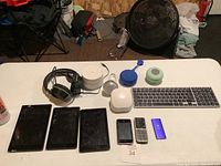 Full view of lot items laid out on table including tablets, headphones, routers, keyboard, phones, iPod, speaker and bulb